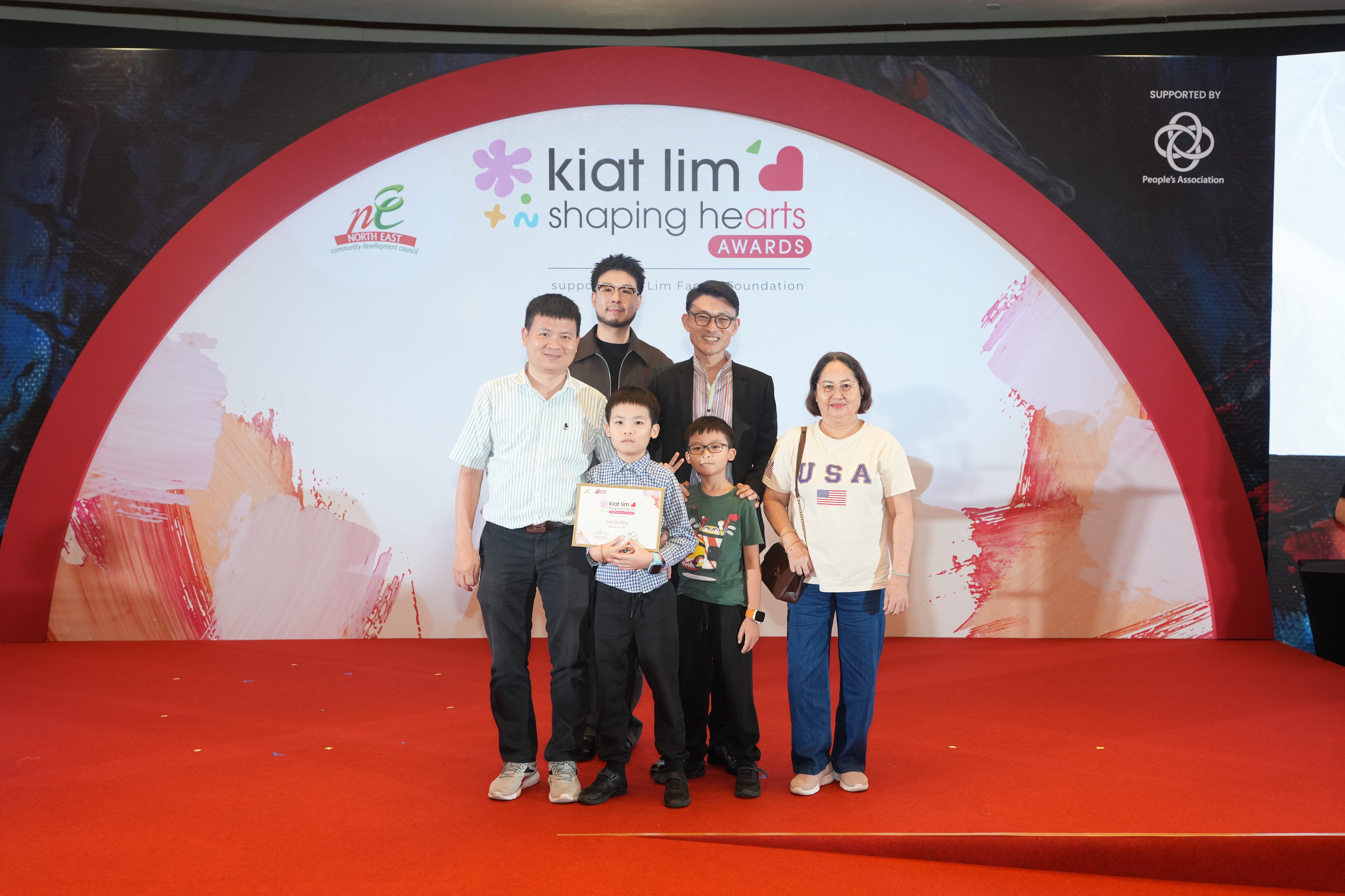 Award recipient standing on stage with the presenter, holding their certificate and posing for a group photo during the award ceremony, with a backdrop reading ‘Kiat Lim-Shaping Hearts Awards'.